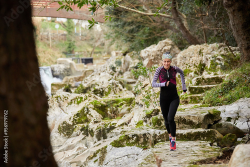 Young trail runner with long blonde hair running along a rocky path next to a river and trees dressed in long tights and long thermal t-shirt in pink, blue and black. She is also wearing a headband. photo