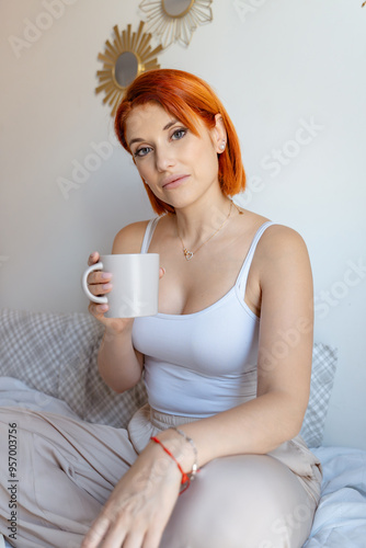 Red-haired woman sits on a bed, holding a coffee cup and looking calmly at the camera. She is dressed in a white tank top and beige pants,