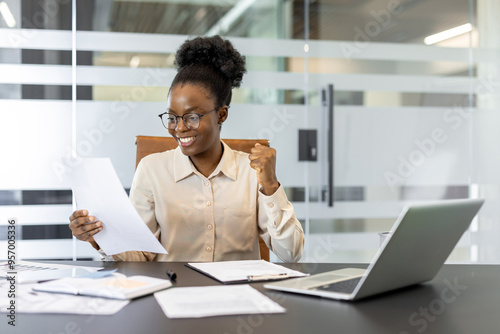 African American businesswoman rejoices at positive news while reviewing paperwork at desk. Bright smile and enthusiastic gesture signify achievement and progress in professional setting