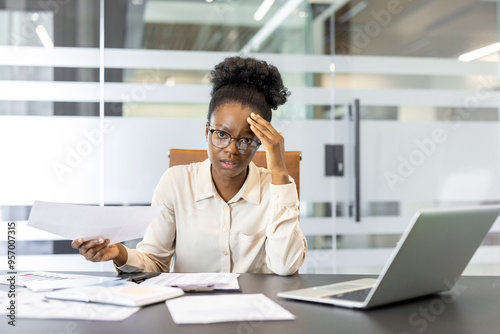 African American businesswoman appears stressed while working at desk with papers and laptop. She holds document and glasses, portraying workplace challenge and professional stress in office setting.