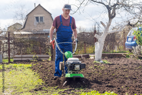 A farmer in the garden tills the land with a motorized cultivator or power tiller, preparing the soil for planting crops
