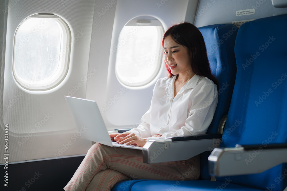 Businesswoman on Flight: A young woman in a white blouse and pink pants is seated on an airplane, smiling as she works on her laptop. The photo captures the sense of calm and productivity that many b Businesswoman on Flight: A young woman in a white blouse and pink pants is seated on an airplane, smiling as she works on her laptop. The photo captures the sense of calm and productivity that many b