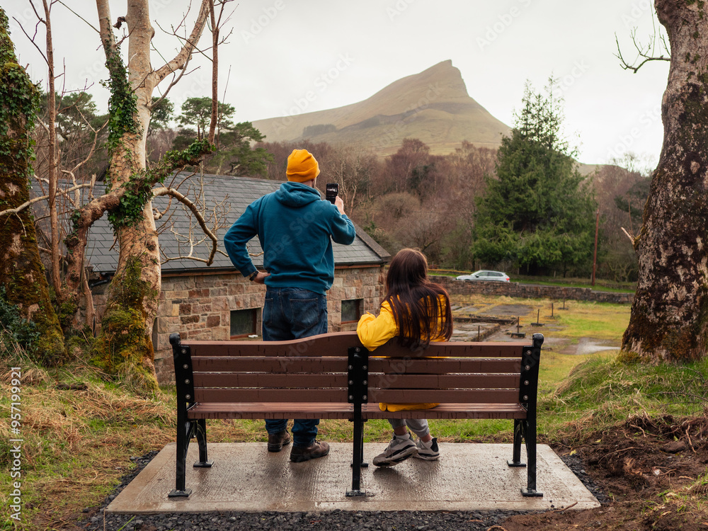 Father and teenager daughter by a bench on a trip. Man taking picture on his smartphone. Nature scenery with road and mountain in the background Divorced family concept.