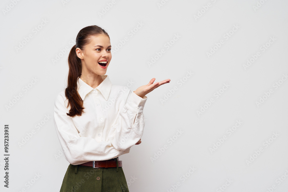 Young woman smiling and gesturing with her hand on a plain white background, dressed in a stylish white shirt and green pants, representing positivity and engagement Young woman smiling and gesturing with her hand on a plain white background, dressed in a stylish white shirt and green pants, representing positivity and engagement