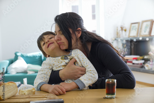 Mother lovingly kisses her son on the cheek while holding him at the kitchen table. Bread and a glass of drink are visible, adding a cozy touch to the heartwarming scene.