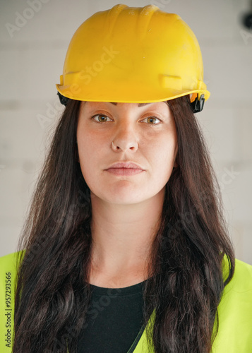Young woman in yellow hard hat and green high visibility vest, long dark hair, looking into camera. Blurred construction site wall background