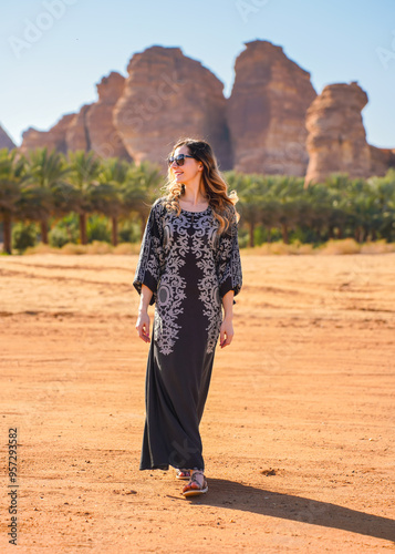 Young woman wearing long dark dress, walking on desert landscape, palm trees and large rock formation typical for Al Ula region in distance. View from behind