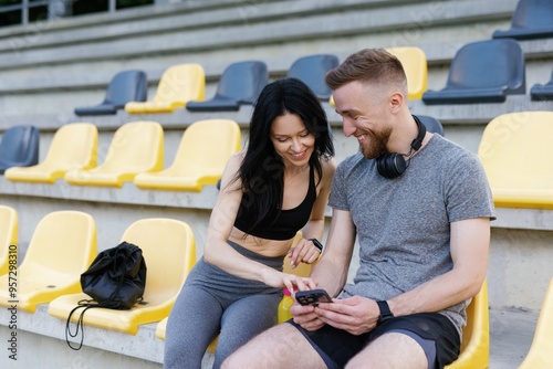Smiling fitness couple relaxing on stadium bench with smartphone