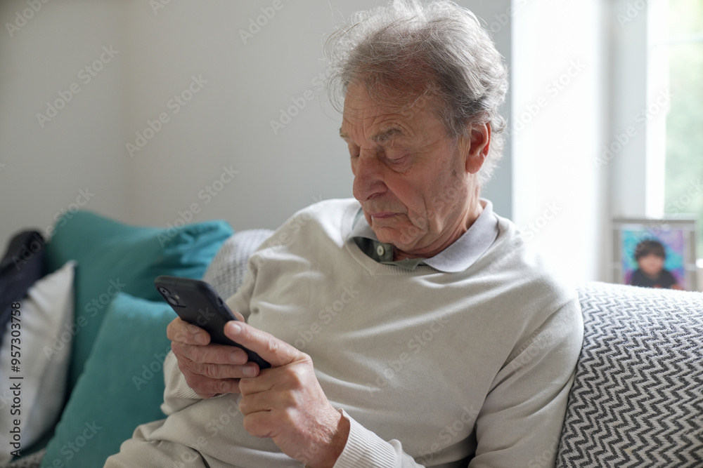 Grandfather sitting on a couch, focused on his smartphone, wearing a light sweater in a cozy living room with cushions and family photos around, representing modern family life and technology use. Grandfather sitting on a couch, focused on his smartphone, wearing a light sweater in a cozy living room with cushions and family photos around, representing modern family life and technology use.