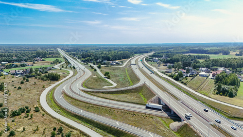 Aerial photograph of a highway intersection with natural, subdued colors. The scene highlights the blend of infrastructure and nature, with roads weaving through a tranquil suburban landscape.