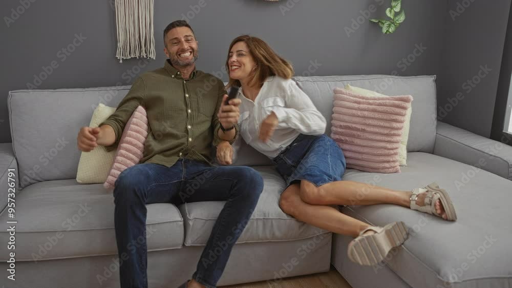Couple laughing on a couch in an indoor living room, holding a remote control and sharing a happy moment together Couple laughing on a couch in an indoor living room, holding a remote control and sharing a happy moment together