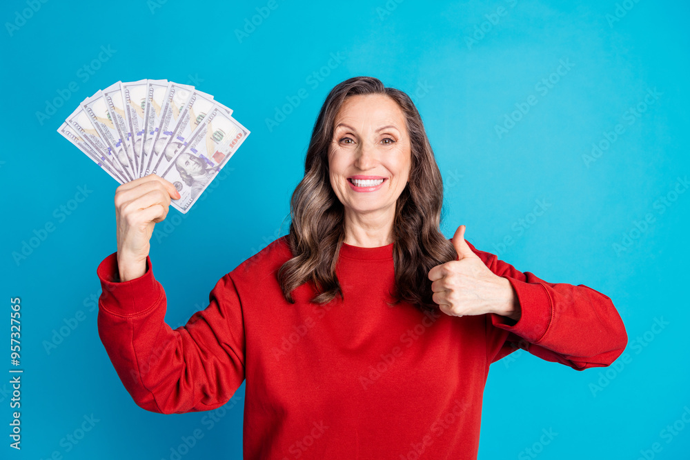 Photo of pretty aged lady thumb up dollar bills wear red pullover isolated on blue color background Photo of pretty aged lady thumb up dollar bills wear red pullover isolated on blue color background