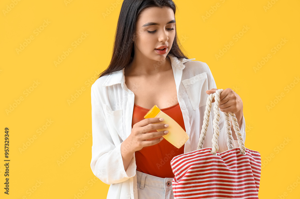 Young woman putting sunscreen cream in beach bag on yellow background Young woman putting sunscreen cream in beach bag on yellow background
