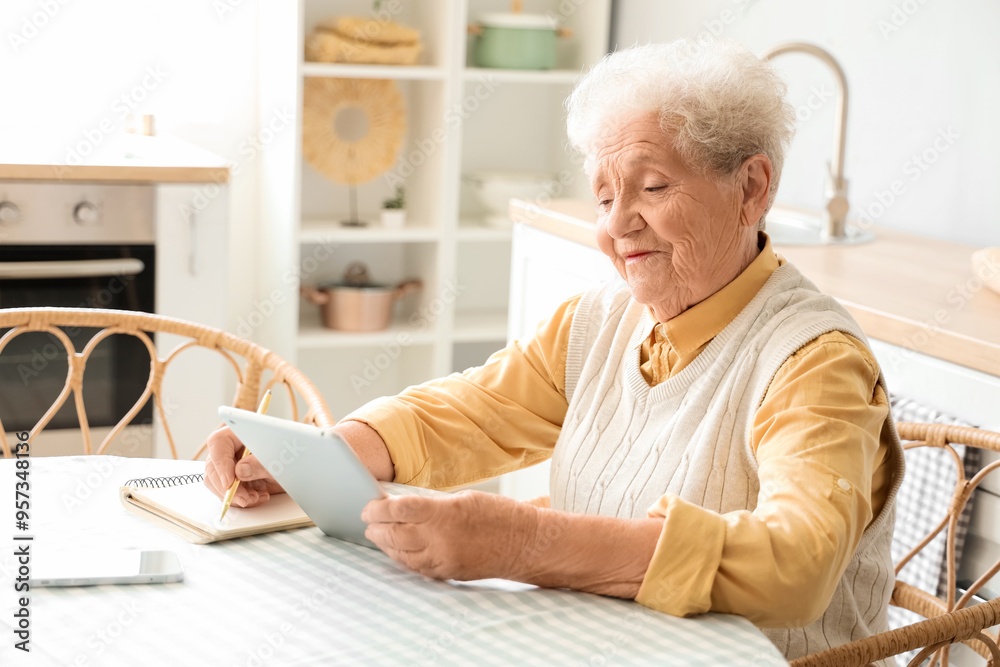 Senior woman with tablet computer writing at table in kitchen Senior woman with tablet computer writing at table in kitchen