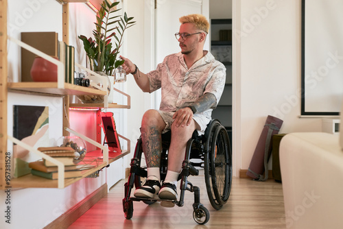 Caucasian man in wheelchair moving through modern apartment reaching for items on shelf. Living room with wooden floor and stylish decor creating a welcoming environment photo