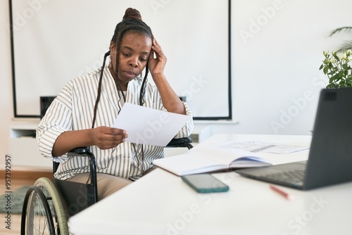 African American woman sitting in wheelchair at work desk, holding document while thinking, with open notebook, laptop, and phone on desk, whiteboard in background photo