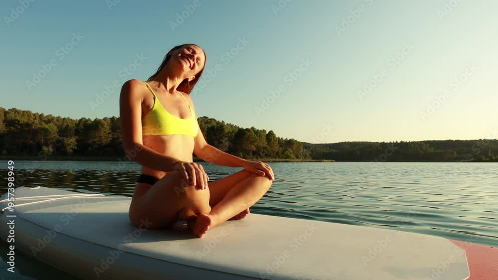 Young woman in bikini performs yoga poses on paddle board, floating serenely on calm lake.  scene captures essence of relaxation and wellness in nature