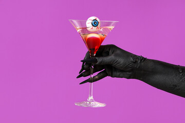 Black hand of witch with glass of cocktail and eye on purple background. Halloween celebration