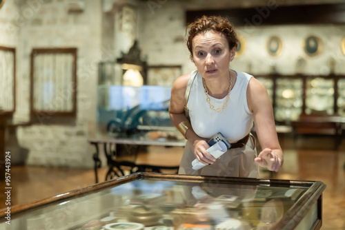 Middle aged woman, wearing white sleeveless top and gold accessories, leaning forward to closely inspect items displayed in glass showcase at museum, demonstrating deep interest in history photo