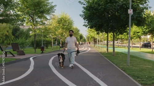 A young man walks his Bernese Mountain Dog on a paved path in an urban park, enjoying a sunny day. photo