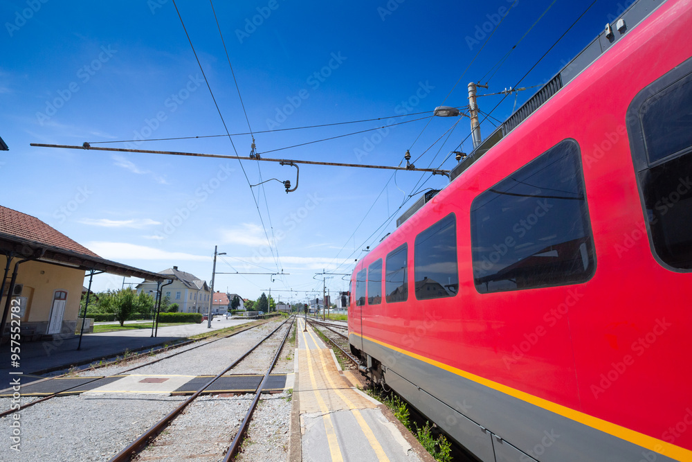 Naklejka premium Slovenian electric train entering Skofja Loka station on a clear day. The modern red train reflects the robust public transportation infrastructure of Slovenia.