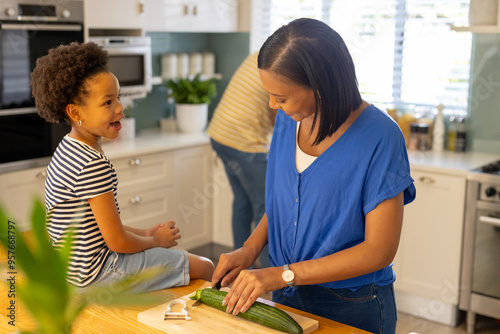 Mother cutting cucumber in kitchen while daughter smiling and sitting on counter