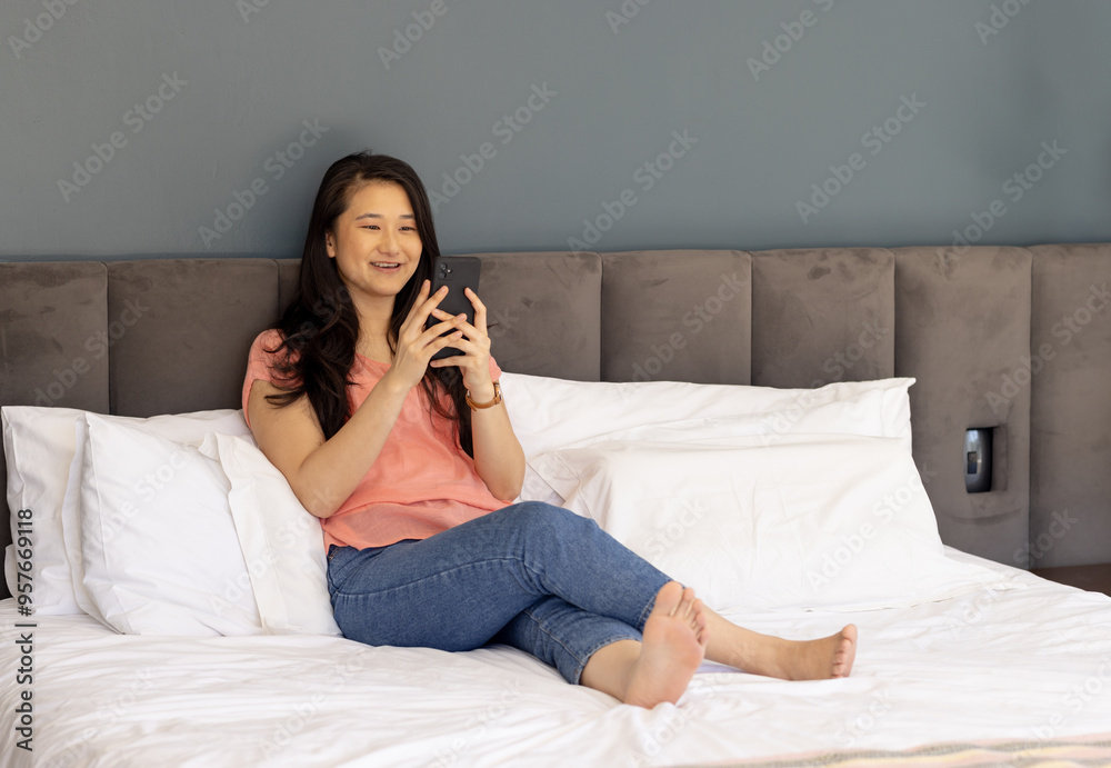 Using smartphone, Asian woman relaxing on bed and smiling in casual outfit Using smartphone, Asian woman relaxing on bed and smiling in casual outfit