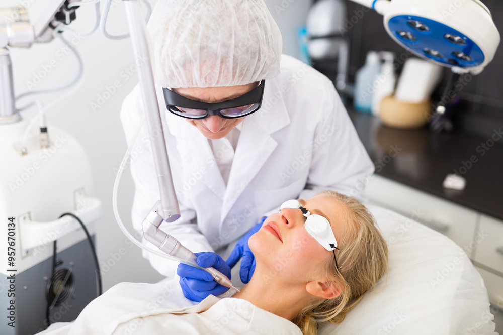 Fototapeta premium Focused skilled female cosmetologist performing hardware laser treatment to resurfacing and tightening facial skin to young woman at cosmetology clinic