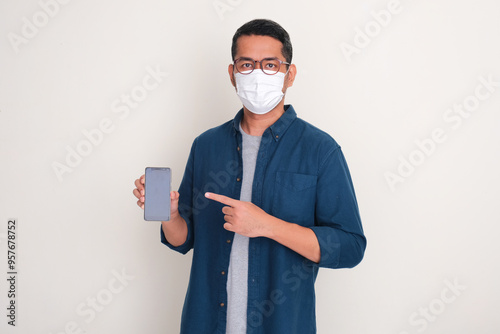 A man wearing medical face mask pointing to blank phone display that he hold photo