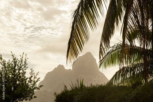 Mount Otemanu on tropical island Bora Bora seen in the distance. Looking through palm tree fronds on a cloudy day in paradise. 