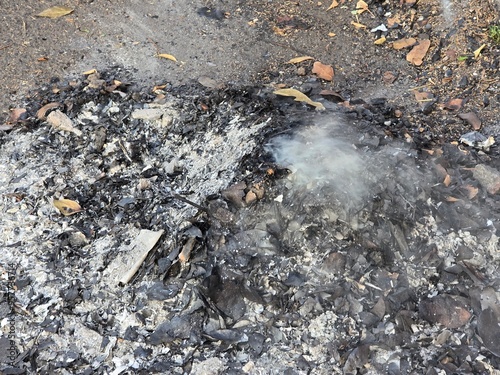 A smoldering pile of ash and charred debris with small wisps of smoke rising, indicating recent burning