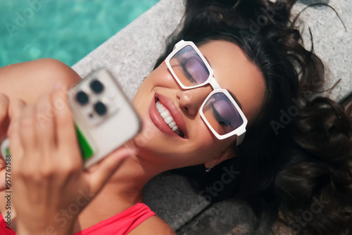 Beautiful girl in pink swimsuit lying on the edge of the pool with smartphone in hand. Tourist using mobile phone in the pool. Relax in luxury pool
