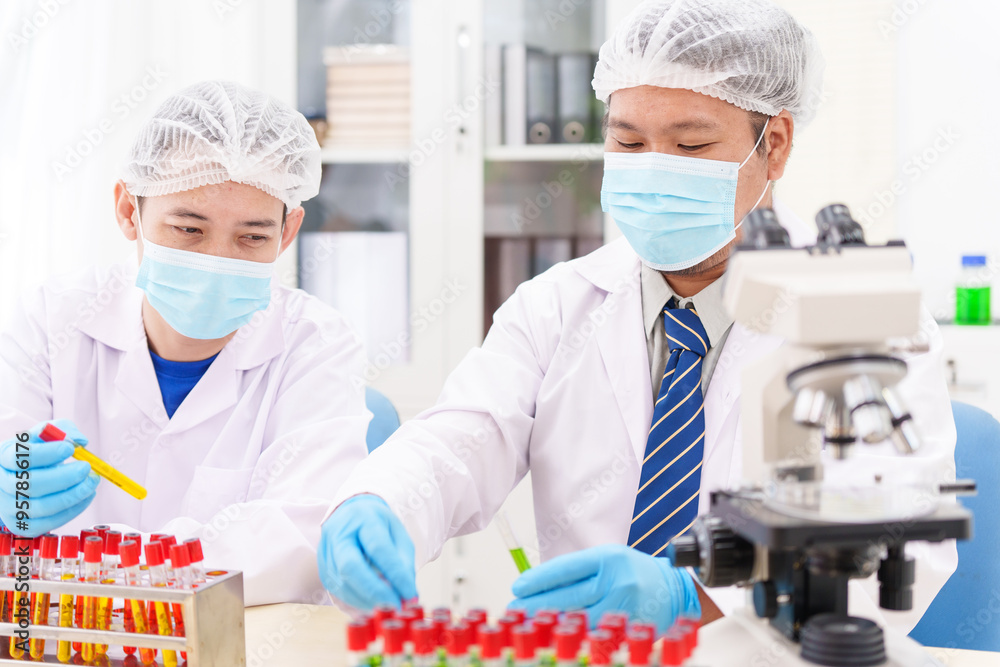Two Asian scientists in lab coats sit at a table in a laboratory, examining samples through a microscope. The room is equipped with scientific tools, liquids, and chemistry materials. Two Asian scientists in lab coats sit at a table in a laboratory, examining samples through a microscope. The room is equipped with scientific tools, liquids, and chemistry materials.