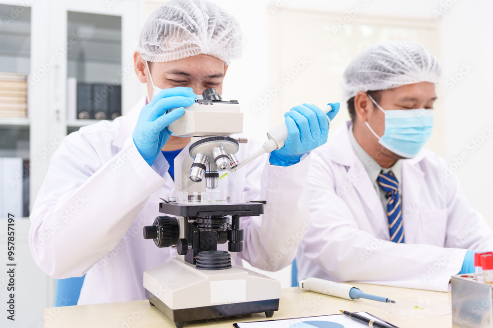 Two Asian scientists in lab coats sit at a table in a laboratory, examining samples through a microscope. The room is equipped with scientific tools, liquids, and chemistry materials. Two Asian scientists in lab coats sit at a table in a laboratory, examining samples through a microscope. The room is equipped with scientific tools, liquids, and chemistry materials.