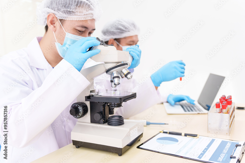 Two Asian scientists in lab coats sit at a table in a laboratory, examining samples through a microscope. The room is equipped with scientific tools, liquids, and chemistry materials. Two Asian scientists in lab coats sit at a table in a laboratory, examining samples through a microscope. The room is equipped with scientific tools, liquids, and chemistry materials.