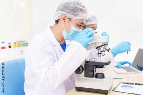 Two Asian scientists in lab coats sit at a table in a laboratory, examining samples through a microscope. The room is equipped with scientific tools, liquids, and chemistry materials.