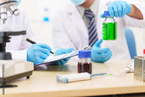 Two Asian scientists in lab coats sit at a table in a laboratory, examining samples through a microscope. The room is equipped with scientific tools, liquids, and chemistry materials.