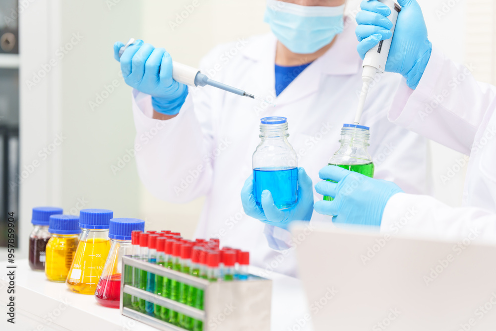 Two Asian scientists in lab coats sit at a table in a laboratory, examining samples through a microscope. The room is equipped with scientific tools, liquids, and chemistry materials. Two Asian scientists in lab coats sit at a table in a laboratory, examining samples through a microscope. The room is equipped with scientific tools, liquids, and chemistry materials.