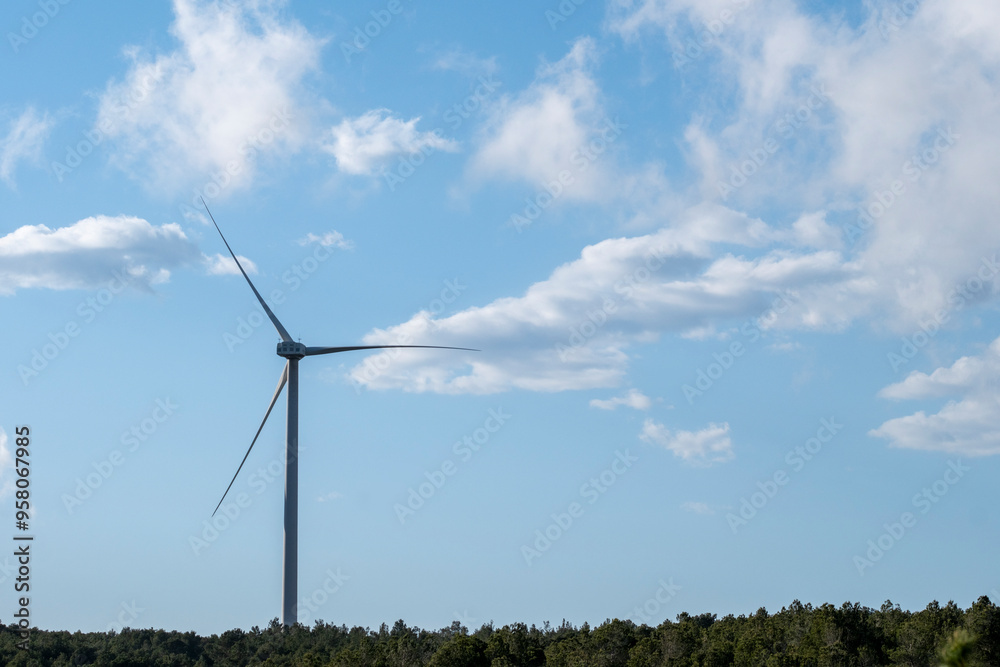 A single wind turbine stands tall under a blue sky with scattered clouds, showcasing renewable energy in harmony with the natural landscape in Tarragona Spain A single wind turbine stands tall under a blue sky with scattered clouds, showcasing renewable energy in harmony with the natural landscape in Tarragona Spain