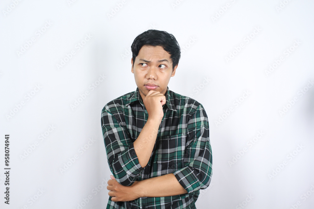 portrait of curious indonesian young man thinking, wondering with hand touching his chin wearing casual green plaid shirt isolated portrait of curious indonesian young man thinking, wondering with hand touching his chin wearing casual green plaid shirt isolated
