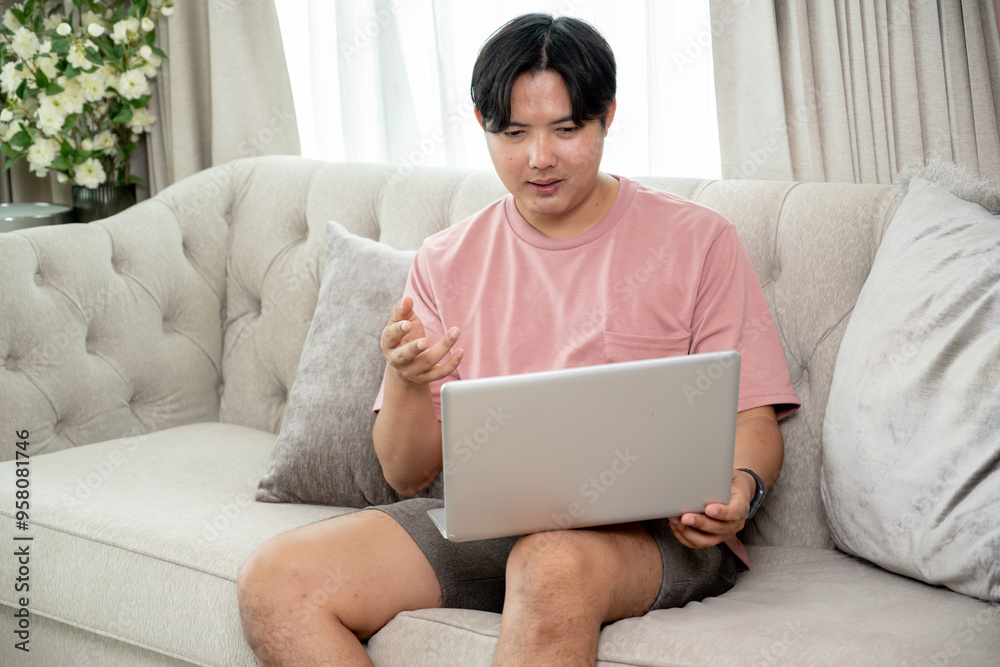 An Asian man in casual clothes using his laptop on a couch in the living room, working remotely from home or scrolling on website.