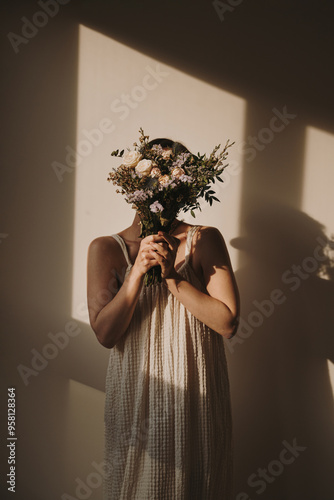 Delicate woman in a light dress holds a bouquet, her face gently hidden. Sunlight and shadows interplay, creating a warm, serene indoor scene. Aesthetic, fashion, and lifestyle vibes