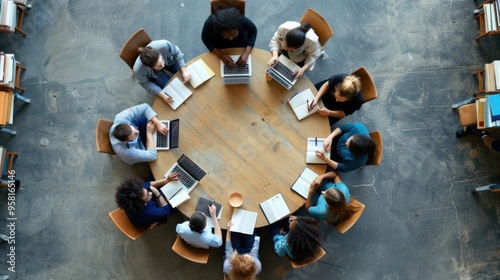 A diverse group of individuals engaged in a brainstorming session around a round wooden table, each using laptops and notepads for collaboration in a well-lit office space