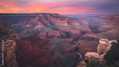 A photo of the Grand Canyon at sunset, with the sky painted in shades of orange and purple. The canyon has a mix of rocky terrain and vegetation. The Colorado River snakes through the canyon. 