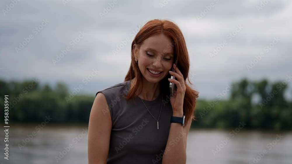 Portrait of cheerful young woman happily chatting on mobile phone while enjoying walk along riverbank, taking in fresh air and scenic views. Smiling redhead female woman talking on smartphone by river Portrait of cheerful young woman happily chatting on mobile phone while enjoying walk along riverbank, taking in fresh air and scenic views. Smiling redhead female woman talking on smartphone by river