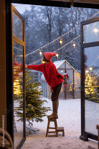 Young woman in red decorates lush Christmas tree with festive ballls and garland at backyard of her house on snow fall, preparing for a winter holidays photo