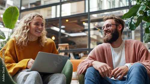 Two people sitting in a modern workspace, engaged in a cheerful conversation while one uses a laptop.