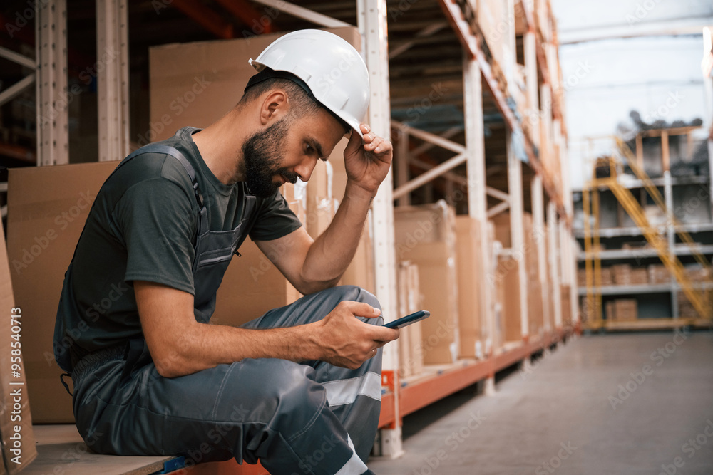 Taking a break, sitting with smartphone. Foreman is in the storehouse, shipping distribution business Taking a break, sitting with smartphone. Foreman is in the storehouse, shipping distribution business