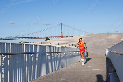 woman jogging outdoors near a modern, open space with a clear blue sky.  She is running is dressed in bright, sporty orange sports bra and pink shorts white sneakers and a smartwatch.