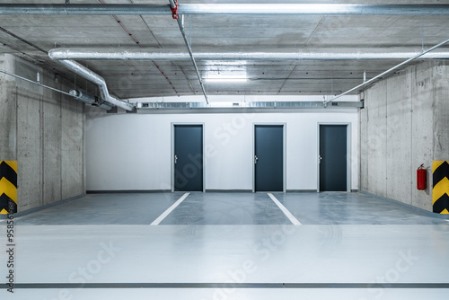 Three black doors are aligned against a white wall in a clean, empty underground garage with smooth concrete floors, bright lighting, and visible utility pipes overhead.